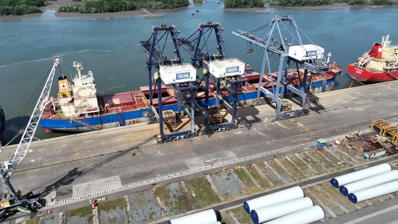 Aerial view of cranes working a ship at the berth, with large steel sections on the quay