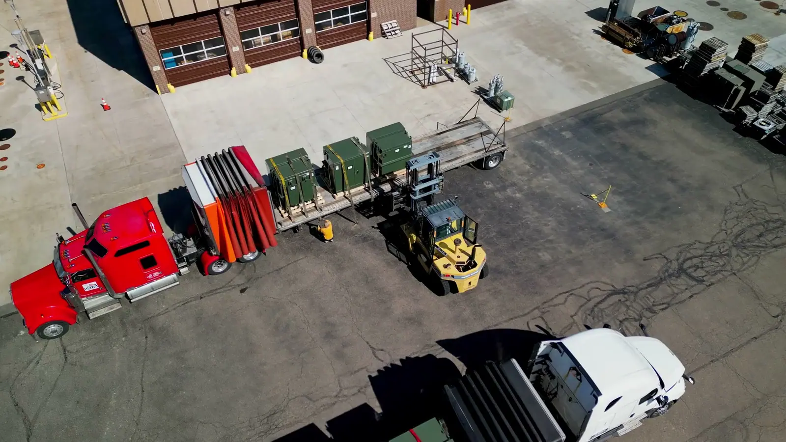 Aerial view of a forklift loading equipment onto flatbed trucks in an industrial yard